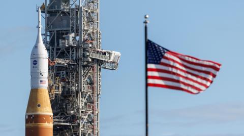 Orange and white Artemis rocket next to a towering grey space system and a blue, white, and red starts and stripes flag, against a blue sky 