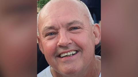 A closeup of a middle-aged man smiling. He has close-cropped grey hair and is wearing a white round-necked T-shirt.