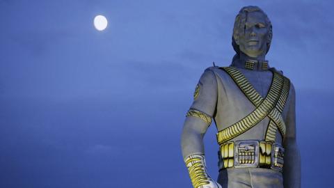 This image features a dramatic nighttime scene dominated by a statue of a Michael Jackson in a highly stylized, futuristic military uniform. 
The uniform is rich with detail—featuring a high collar, ornate shoulder pads, and metallic bands that crisscross the chest and arms. The statue exudes a commanding, almost regal presence, enhanced by the intricate textures and utility-style belt with multiple compartments.

Behind the figure, a full moon glows in the upper left corner, casting a serene but stark light that heightens the statue’s contours