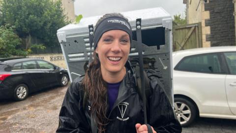 Dee Woodwaid in a rain proof jacket with a fridge strapped to her back. She is laughing and looking at the camera.