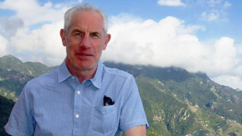 A man with white hair wearing a blue short sleeved shirt against a backdrop of mountains