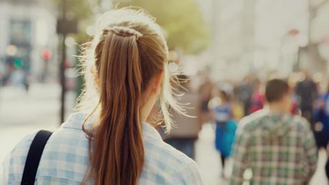 An anonymous young woman with long hair and a blue checkered shirt walks down a street with passers by in the background