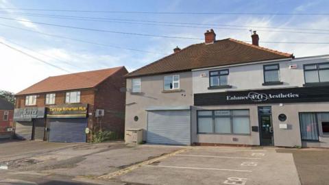 A row of shop units in a residential street. The unit in the centre has metal shutters down and is unused.