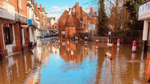 A flooded street in the town centre of Tenbury Wells, with orange and white traffic cones and barriers placed in the waters. Redbrick buildings line the streets.