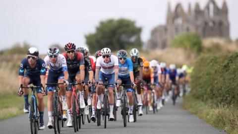 A group of cyclists, wearing colourful Lycra cycle along a paved, single track road, surrounded by hedgerows. In the background, Whitby Abbey, a ruined stone building, can be seen.