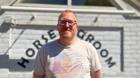 A man wearing glasses and a cream T-shirt with a logo on it smiles towards the camera, while standing outside a white-coloured pub building. 