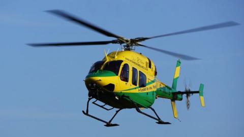 Image of a Wiltshire Air Ambulance helicopter in the sky. It is yellow and green.