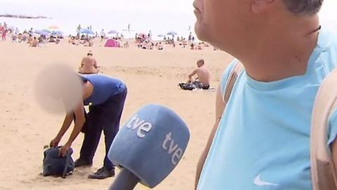A man talking to TV crew while a man steal a bag on the beach