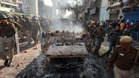 Police walk past a vehicle set on fire by protesters in Delhi