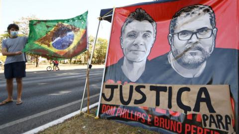 A man holds a Brazilian flag tinted with red next to a banner in demand of justice for the killing of British journalist Dom Phillips and Brazilian indigenist Bruno Pereira
