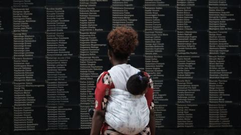 A woman carrying her child looks at a memorial to victims of the 1994 Rwandan Genocide