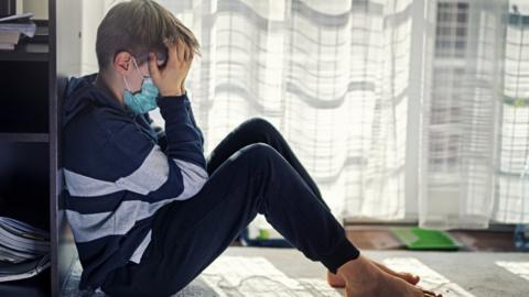 A boy with his face in his hands, wearing a mask