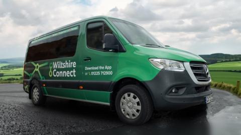 A green mini bus with Wiltshire Connect written on the side, on a road in the Wiltshire countryside, green hills behind.