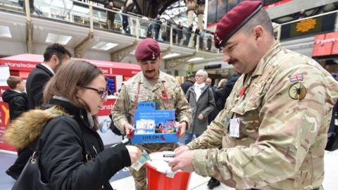 A young woman puts money into a collection bucket held by a veteran in uniform