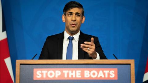 Rishi Sunak stands at a podium and gestures with a slogan "Stop the Boats" written beneath him