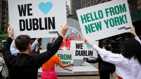People hold signs as they greet during the reveal of The Portal, a public technology sculpture that links with direct connection between Dublin, Ireland and the Flatiron district in Manhattan, in New York City, U.S., May 8, 2024.