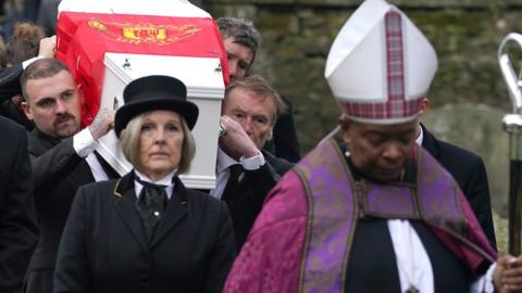 William's white coffin wrapped in a Manchester United flag, carried by people in black suits and led by the bishop