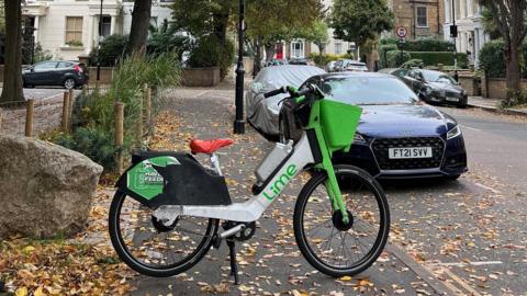 An ebike is parked across the pavement. There is a car in the distance.