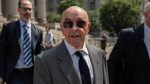 Joe Lewis stands outside a courthouse and wears a grey suit with a black tie and sunglasses.