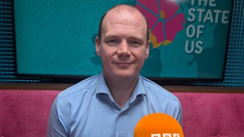 Gordon Lyons. He is bald and wearing a light blue shirt. He is sitting on a pink sofa in front of an orange BBC Sounds microphone. The State of Us branding is on a screen behind him.