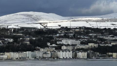 A photos of snow on the hills behind Onchan Head. There are large white buildings on the seafront below and darkening blue sky above.