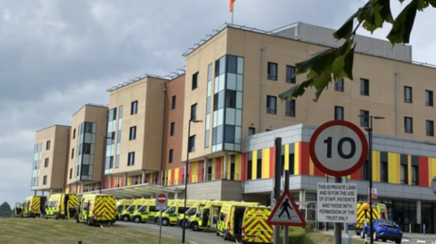 A large, multi-storey beige building with a row of yellow ambulances parked outside