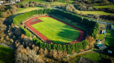 An aerial photo taken of the Whitchurch race track. There is a green field in the middle of the red oval running track and the track is surrounded by thick green trees and bushes.