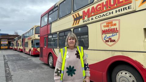 A woman with light brown hair cut into a fringe, wearing a high-vis jacket over a knitted beige jumper with a floral design, looks into the camera on a concrete bus depot with red and yellow double decker buses in the background. 