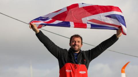 Sam Goodchild, standing outdoors, holding a Union Jack flag above his head with both hands. He has short brown hair and a short beard, and is wearing a red and black top.