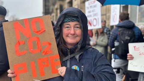 Protester Tina Lancaster wears a navy coat with the hood up and holds a sign which reads 'No Co2 Pipe' in orange writing outside Wallasey Town Hall. 