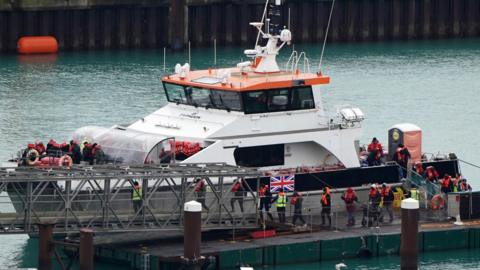 A white boat with an orange roof by the side of a jetty. Several people in dark clothes and lifejackets are disembarking.