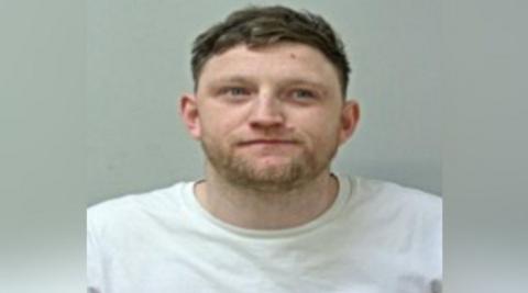 Mugshot of Daniel Burba. He is wearing a white T-shirt and has a stubbly beard and short brown hair