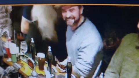 a man with a beard in grey jumper smiling broadly as a he sits at the bar with the beer taps in front of him. He is also holding a mobile phone