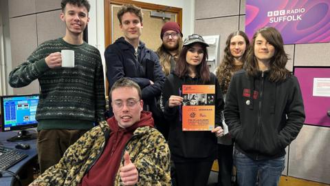 Seven young people pose for a photo in a radio studio. One is sitting down giving a thumbs up signal to the camera. The others are standing a smiling at the camera and one person is holding up an orange poster advertising the gig that the group are hosting. A sign for "BBC Radio Suffolk" is behind the group of people. 