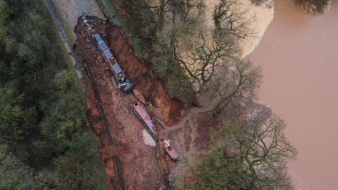 An aerial shot shows a big brown body of water on the right hand side. In the middle, there is a large muddy hole with two red narrowboats at the bottom. They both have blue solar panels on their roofs. The trench has a tree line either side of it.