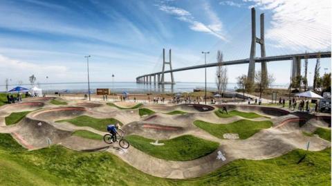 A CGI image of how the pump track could look. It is a looping circuit with a cyclist jumping over one of the humps. The path is surrounded by grass and the sea is in the background.