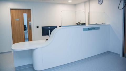 A white reception desk with clear glass screens on top and a clock on the wall. 
