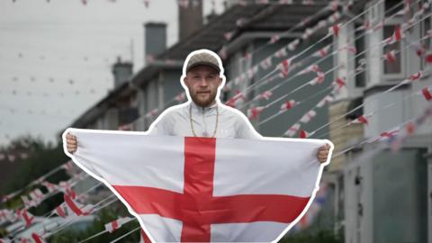 A man stands with an England flag outstretched in his arms. He has a beard, cap and gold chain around his neck. Behind him is a blurred picture of a row of houses with a string of England bunting draped from them. 