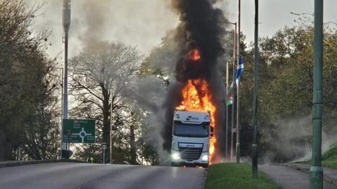 A photo taken from a distance of a lorry on fire on the side of a road. Flames can be seen coming from all sides of the vehicle and thick black smoke billows up into the sky. A pavement and grass runs alongside the road and there is a green roundabout road sign on the left. 