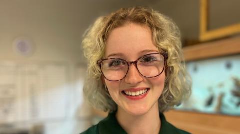 Zoe Cousins smiles broadly at the camera, she is wearing large glasses and has short, blonde curly hair. She is standing in front of a fish tank. 