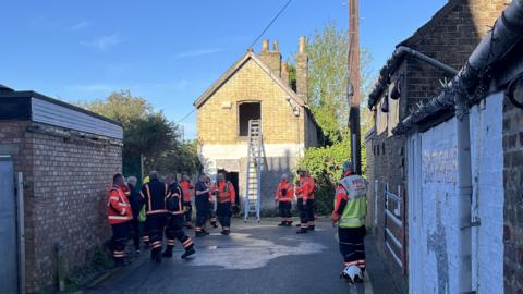 Firefighters are gathered in a lane. There is a ladder against a brick building going up to a first-floor window opening.