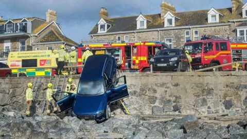 A blue Audi estate car, with its bonnet on breakwater boulders, it rests on a granite wall slightly higher than a grown man, with both front doors open and one hanging off. It is surrounded by firefighters and ambulance crew, as well as emergency service vehicles.