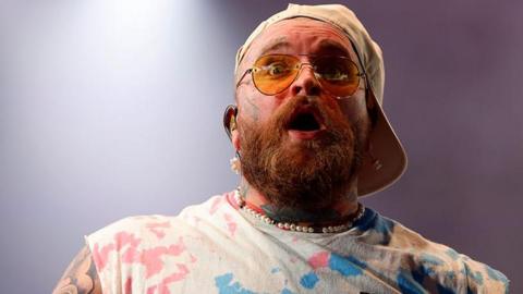 Teddy Swims with a light brown beard wearing a beige baseball cap sideways and sunglasses, pearl necklace and a white tye-dye t-shirt. He is on stage performing and looks surprised with his eyes wide and mouth open facing towards the crowd at Radio 1's Big Weekend festival.