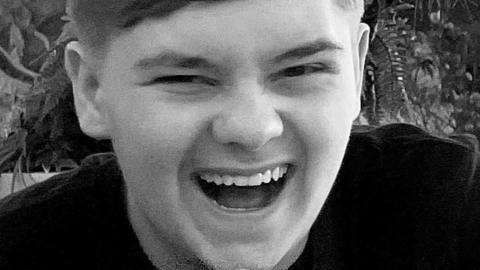 A young man wearing a dark T-shirt looks towards the camera and is seen to be laughing in a black and white photograph.