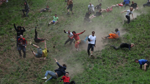 Image shows men running and tumbling down Cooper's Hill. The hill is very steep and grassy. The ground is so dry that there are dust trails behind the men as they run. Mot of the men have fallen over and are rolling and falling down the hill, with only a few of them still on their feet.