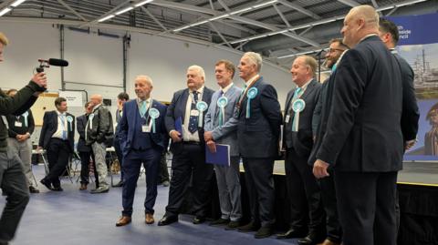 Eight men stand in a line in front of the stage and smile for the photographer in front of them. They are all wearing turquoise rosettes for Reform.
