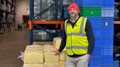 A man wearing a red hair net and hi viz is standing in front of a pallet of cheese in a warehouse, which is full of pallets of cheese.