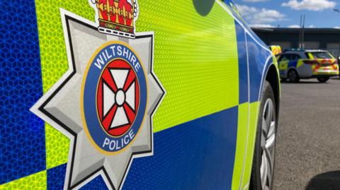 The side of a marked Wiltshire Police car, which is parked in a car park. Another police car is parked in the background. 