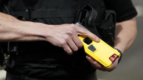 Stock image of an officer demonstrating a new Taser. It is a close up shot of a man's hands holding a yellow taser, with the name Taser 7 on it and there are two red cartridges visible.