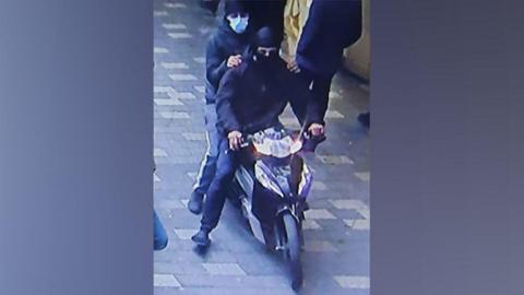 Two men wearing balaclavas and facemasks on a moped riding amongst people.